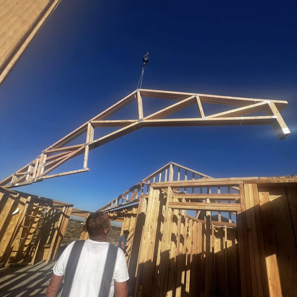 A wood roof truss lifted into place by a crane against a blue sky, with crew below
